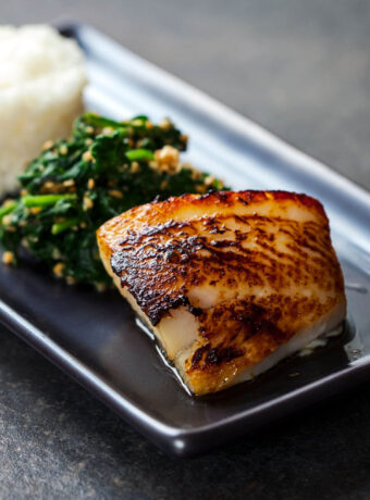 A filet of miso black cod on a black plate served with spinach and white rice. A delicious Japanese dinner.