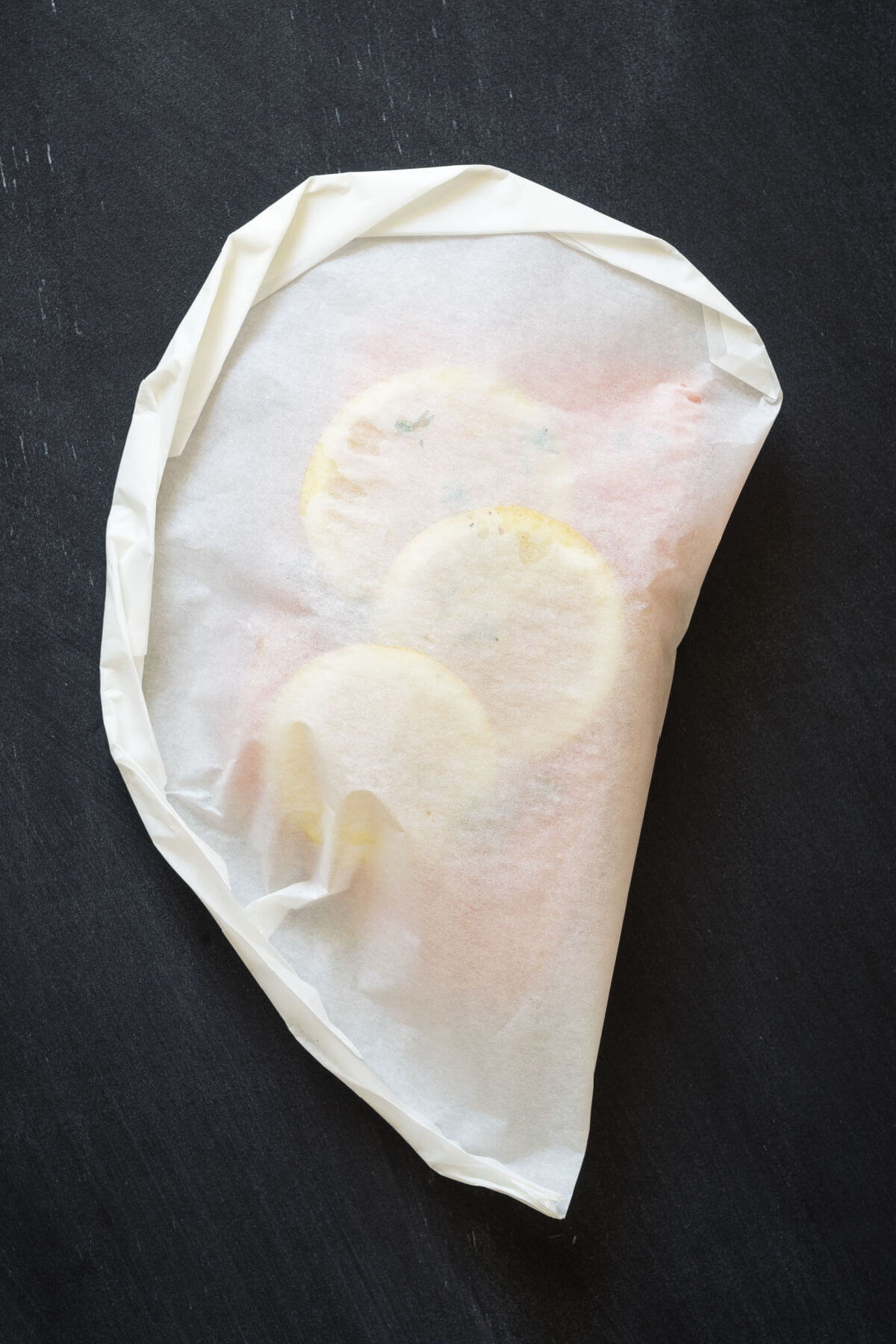 A parchment paper pouch filled with fish, lemons, and herbs on a black background. 