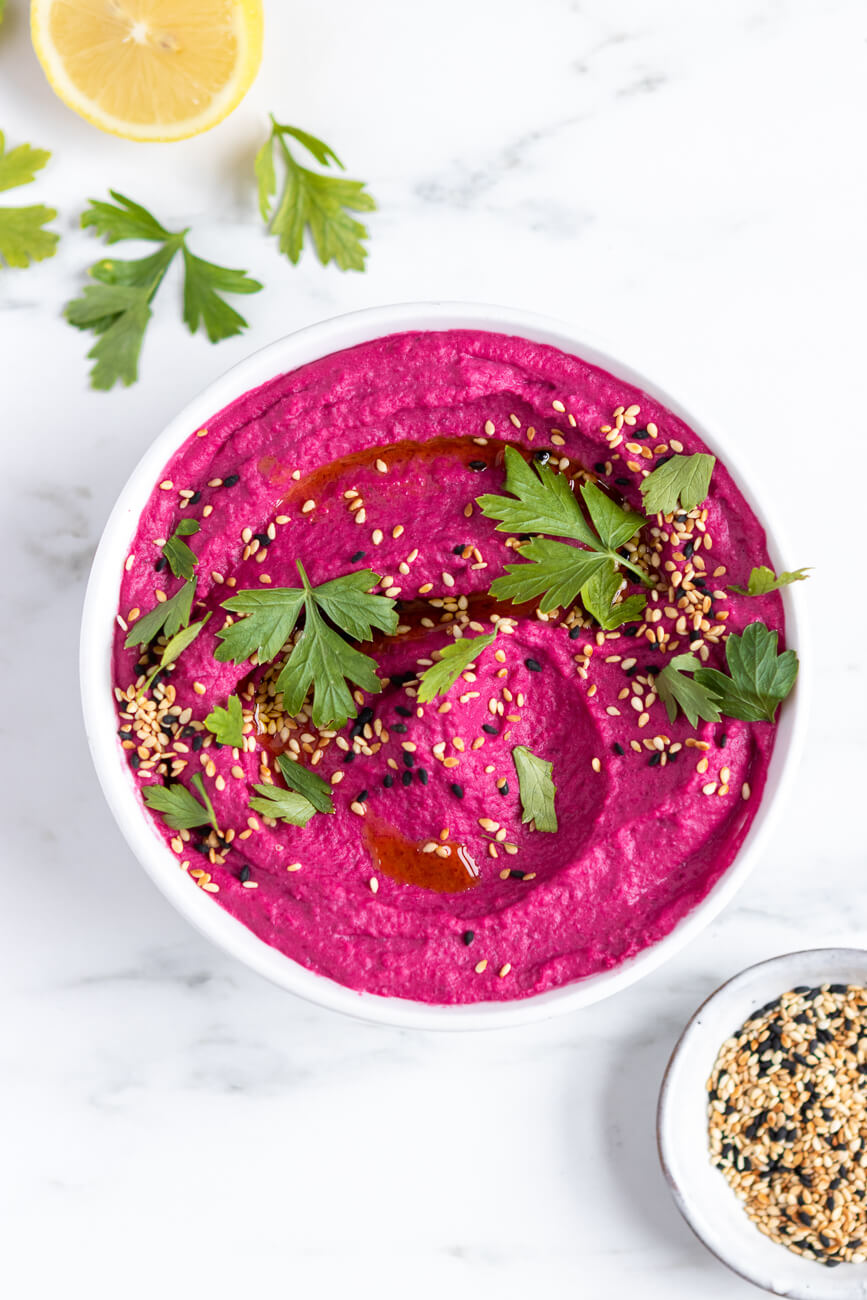 An overhead photo of a white bowl filled with bright pink beet hummus topped with sesame seeds and parsley.