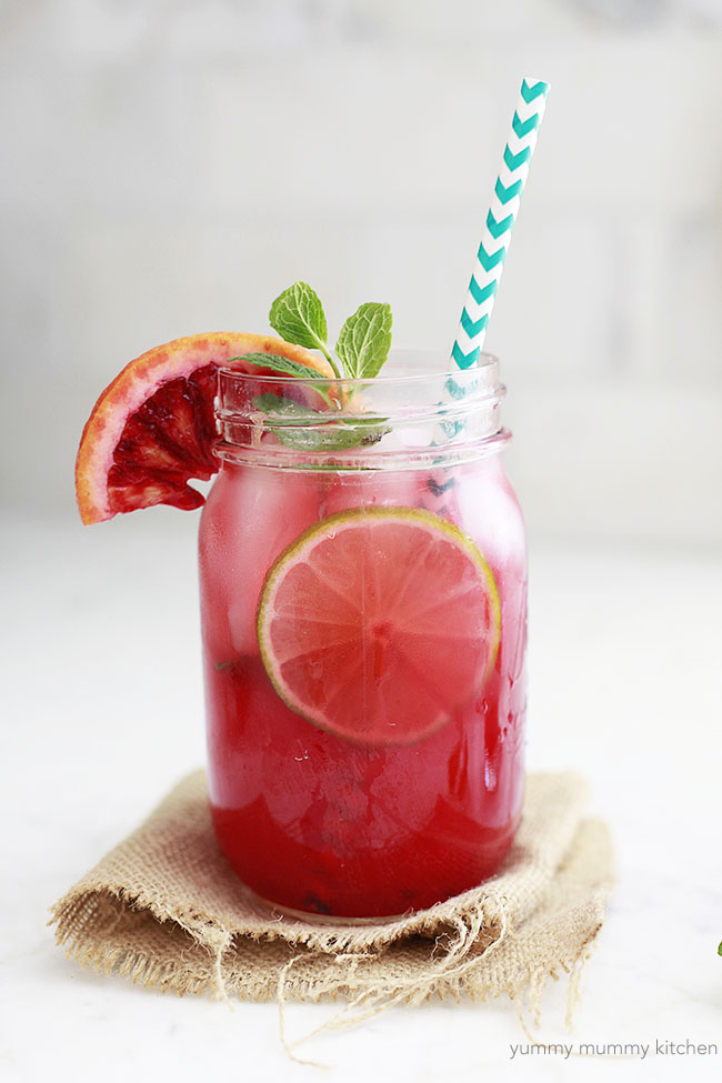 A mason jar filled with a bright pink blood orange margarita garnished with fresh blood orange and lime slices and mint. The drink has a paper straw and sits on a marble kitchen counter. 