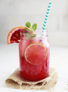 A red blood orange margarita in a mason jar garnished with lime and blood orange slices and a sprig of mint. The glass sits on a white kitchen counter.