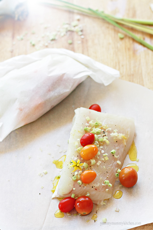 A piece of raw halibut with minced lemongrass and cherry tomatoes on a piece of parchment paper. 