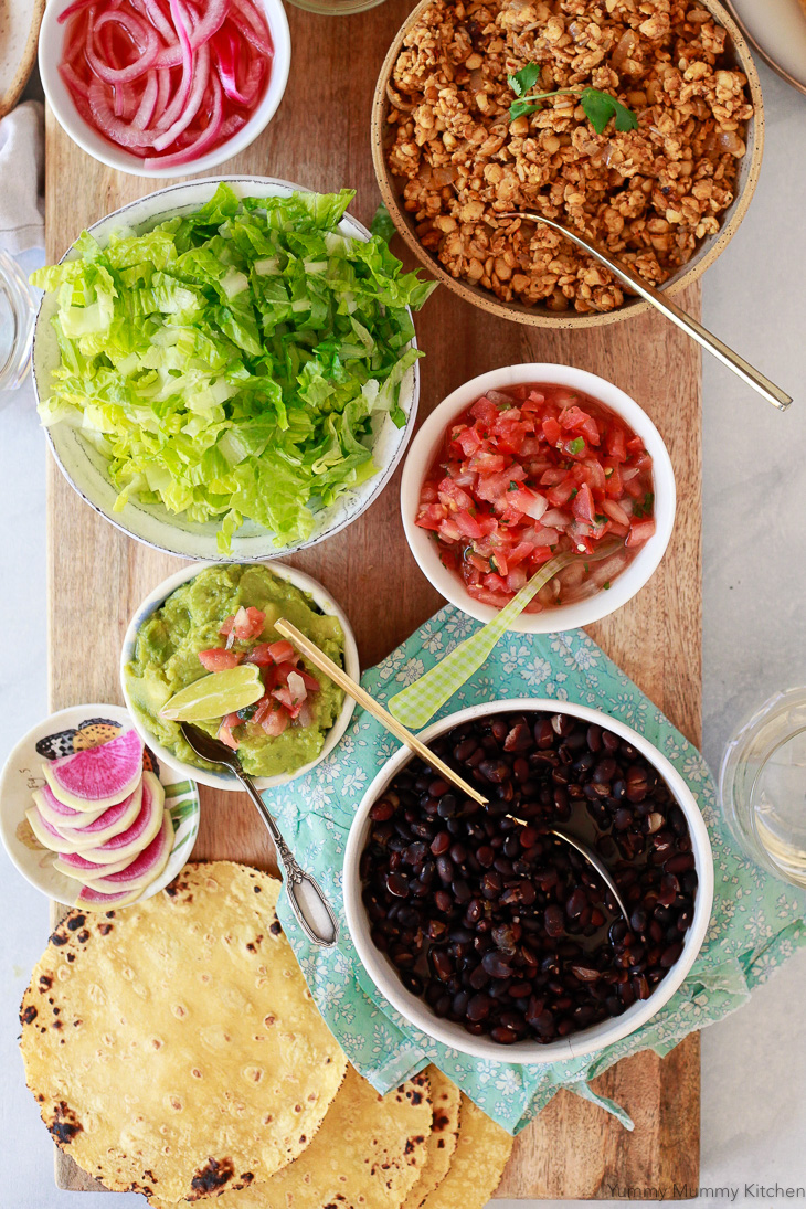 Tempeh taco filling along with black beans, salsa, guacamole, and lettuce on a long serving board. 
