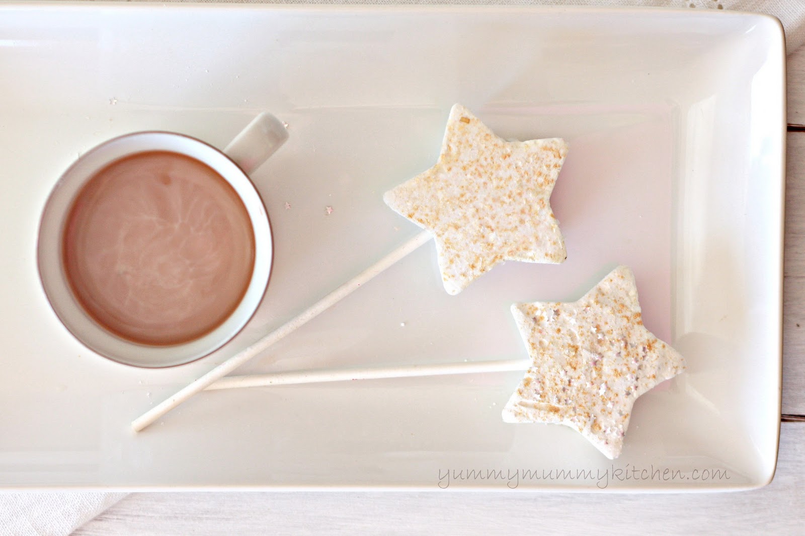 Two star-shaped marshmallows topped with edible gold glitter with sticks for stirring on a whit eplatter next to a mug of hot chocolate. 