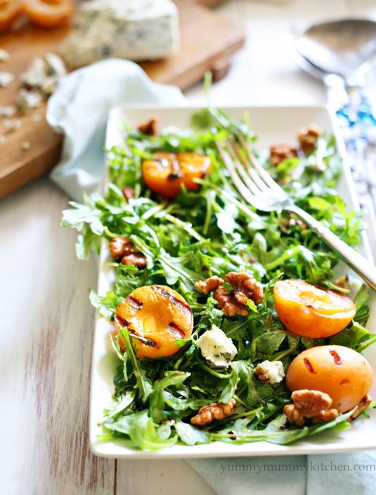 An arugula salad with gorgonzola cheese, walnuts, and grilled apricot halves on a white rectangular plate. 