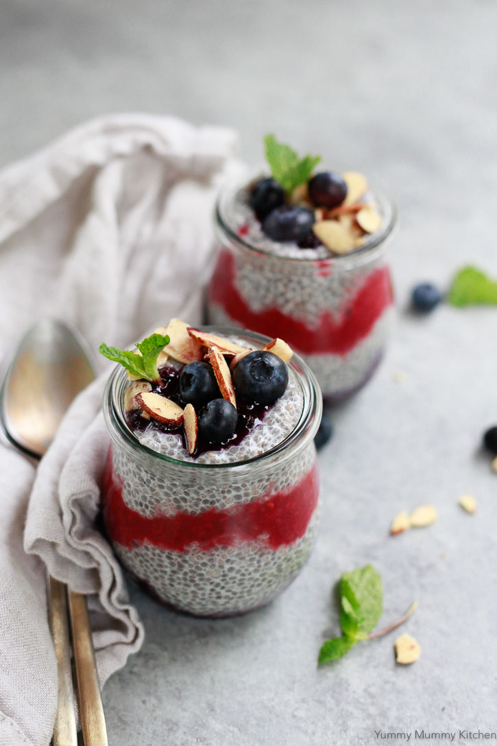 Two small jars filled with chis pudding with berries and berry puree swirls on a concrete surface. 