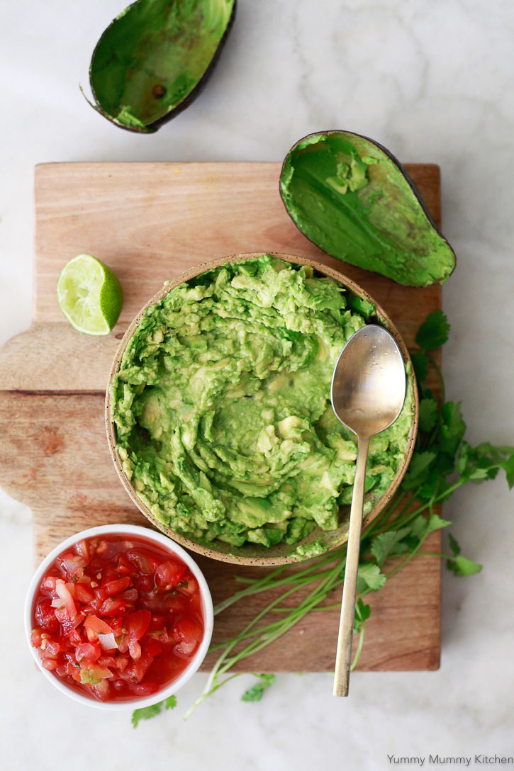 A bowl filled with homemade guacamole on a cutting board next to avocado peels and a cup of salsa. 