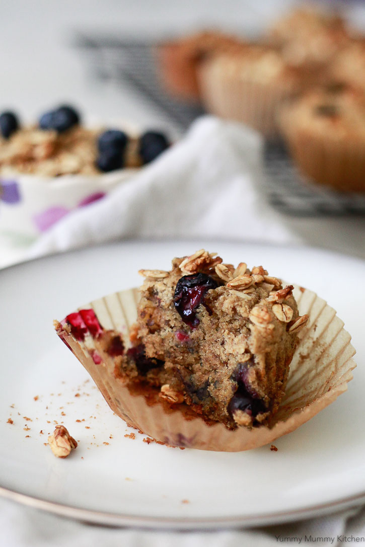 Half of a gluten free vegan banana muffin with granola and blueberries in a parchment baking cup. 