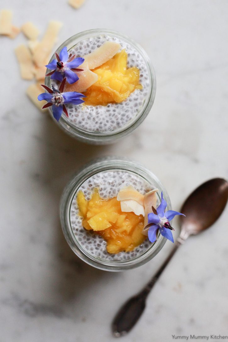 An overhead photo of two small jars filled with coconut milk chia seed pudding topped with mango puree, coconut chips and purple borage edible flowers. 