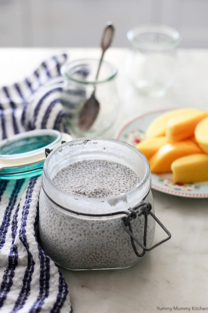 A glass jar is filled with creamy coconut chia seed pudding. A sliced mango on a plate can be seen in the background along with two small serving jars. 
