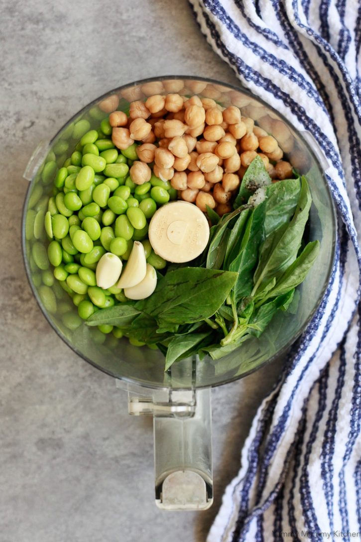 Chickpeas, edamame, basil, and garlic in the bowl of a food processor ready to make homemade edamame hummus. 