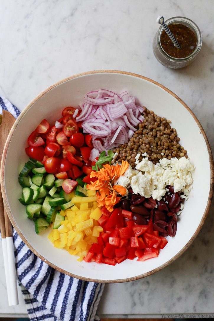 A beautiful Mediterranean lentil salad bowl with cucumber, tomatoes, olives, feta, and bell pepper. Greek salad dressing sits on the side. 