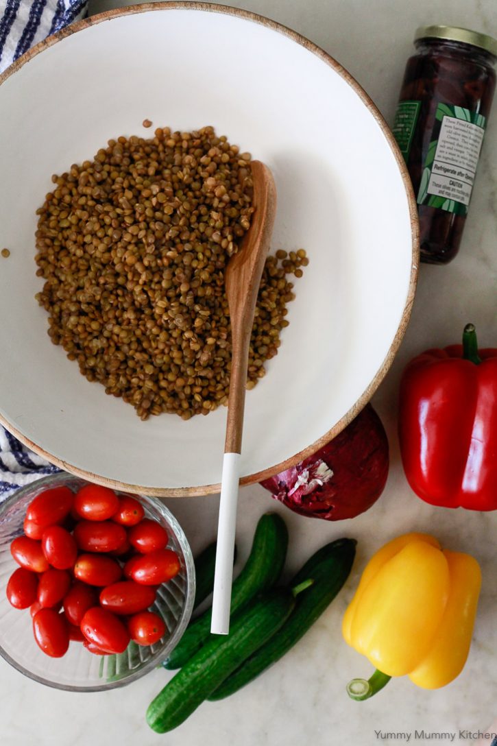 Mediterranean lentil salad ingredients on a white countertop include cooked green lentils, tomatoes, cucumber, bell peppers, and olives. 