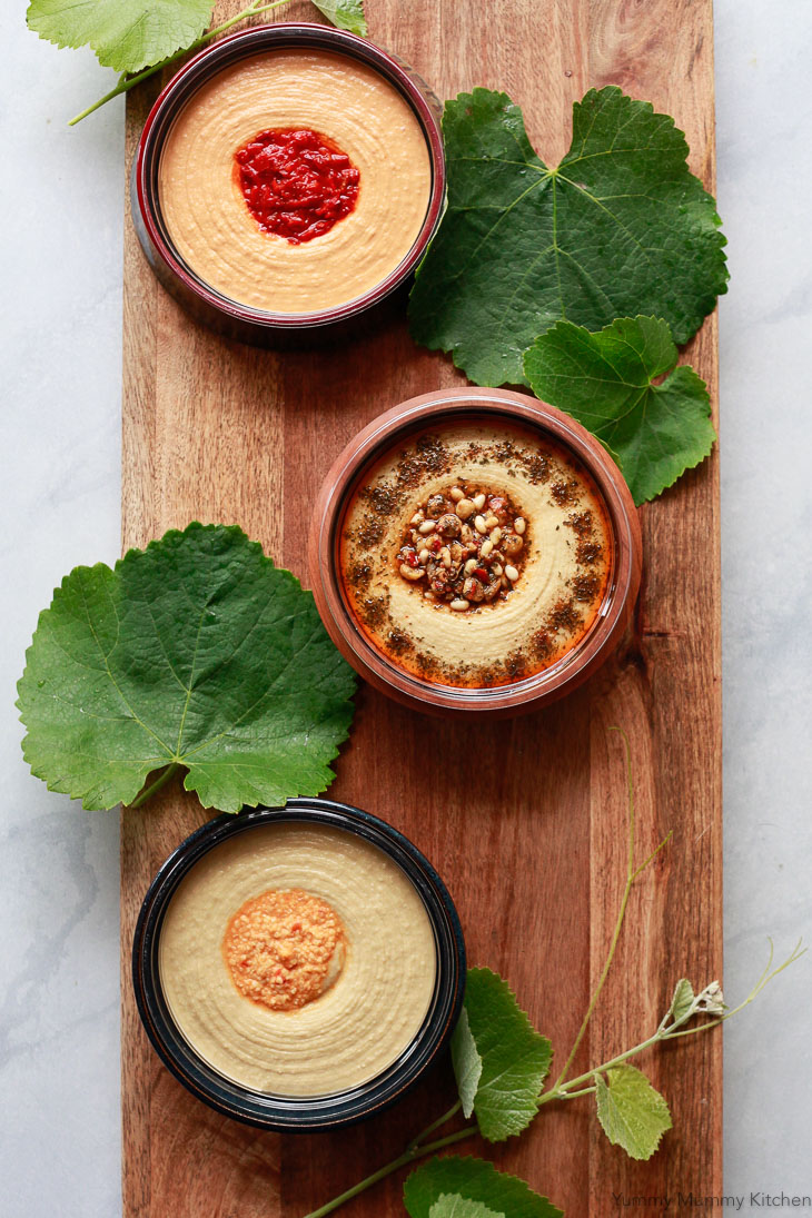 Three Sabra Hummus tubs in handmade bowls on a wooden board with grape leaves are the start of a vegetarian mezze platter. 