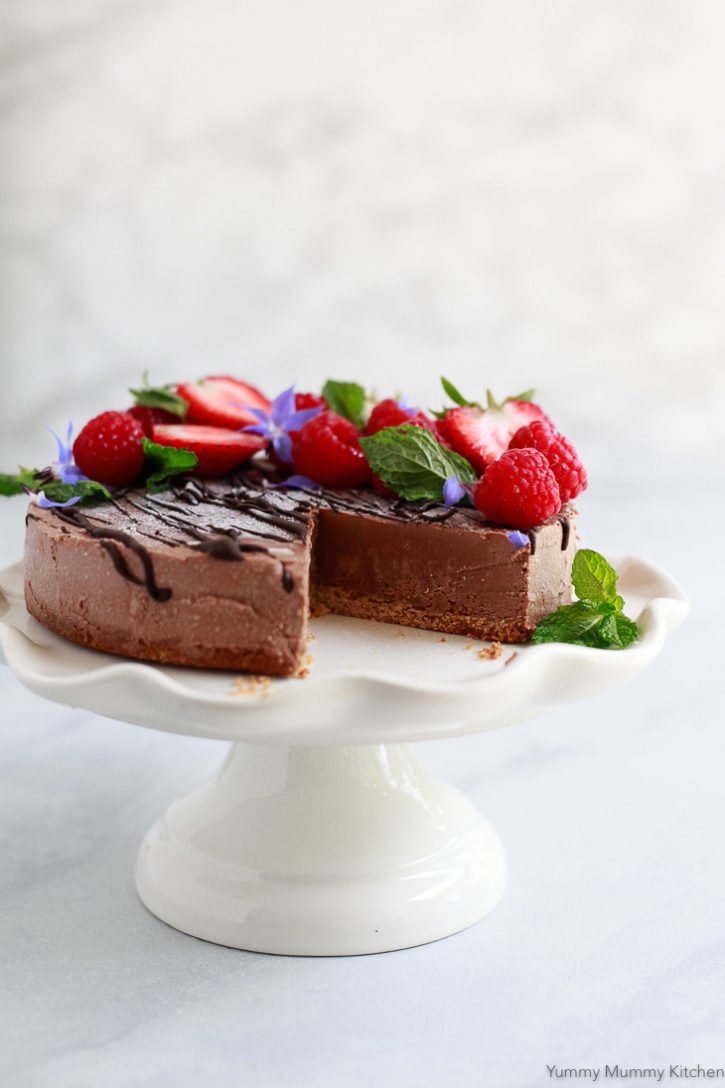 A no-bake vegan chocolate cheesecake on a small white scalloped cake stand. The chocolate vegan cheesecake is topped with fresh berries and mint. 