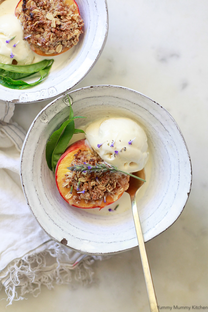 Two white ceramic bowls filled with baked peaches topped with crumble topping served with vanilla ice cream and lavender flowers. 
