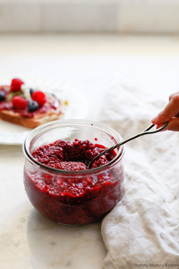 A jar of raspberry chia jam with a jam spoon. This easy raspberry chia jam is a healthy breakfast addition with no added sugar. 