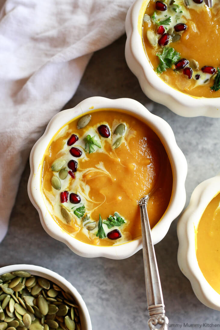 A beautiful bowl of pumpkin soup in a pumpkin-shaped white ceramic bowl. The orange colored soup is topped with swirls of cream, pepitas, herbs, pomegranite arils. 