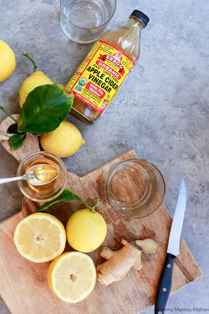 Ingredients for an apple cider vinegar drink recipe including lemons, Bragg ACV, ginger, and honey on a cutting board. 