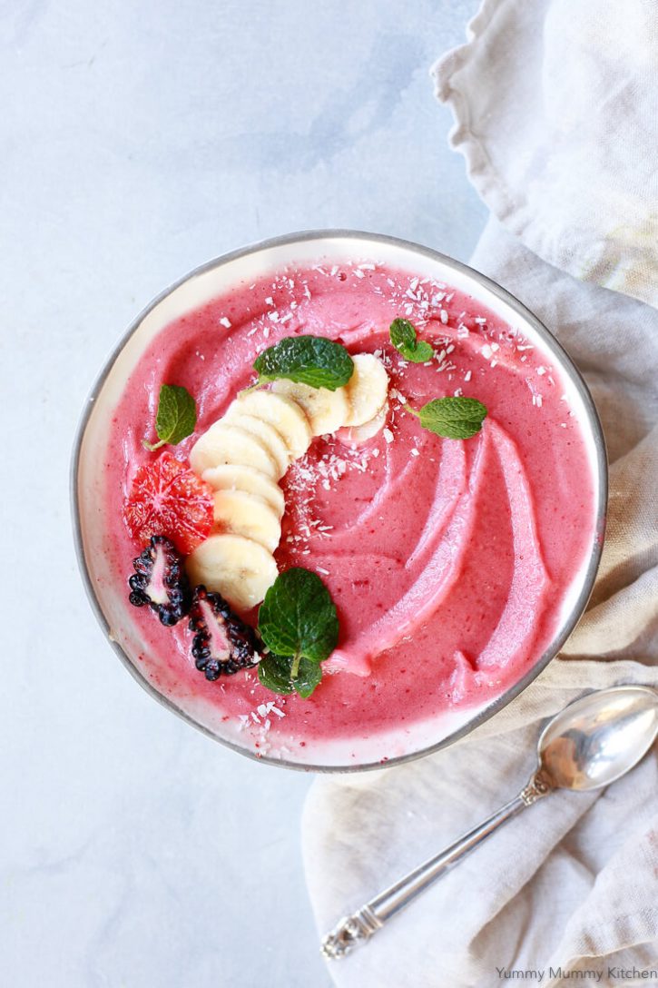 A beautiful smoothie bowl filled with a strawberry banana smoothie and topped with sliced banana, blackberries, orange, and mint leaves with a sprinkle of coconut shreds. 