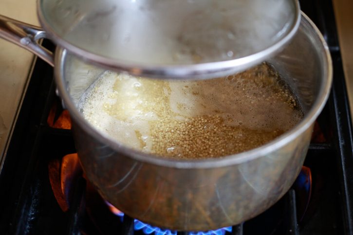 A saucepan filled with quinoa and water is covered with a lid on the stove to cook quinoa. 
