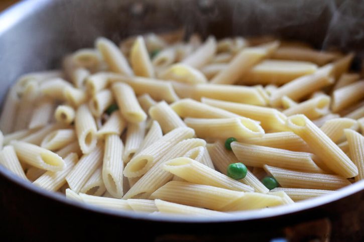 A close-up image of steaming cooked penne pasta with peas in a pot. 