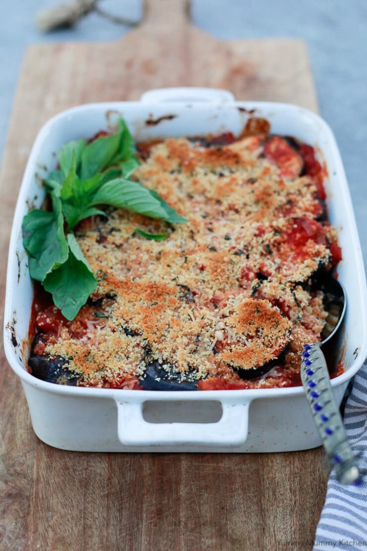 A white casserole dish with filled with baked vegan eggplant parmesan topped with golden brown breadcrumbs and garnished with fresh basil. 