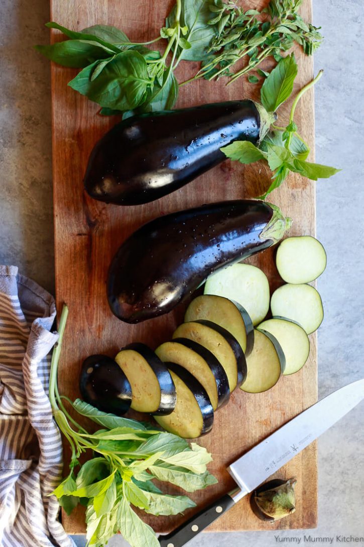 Eggplants are sliced on a wooden cutting board with basil. 