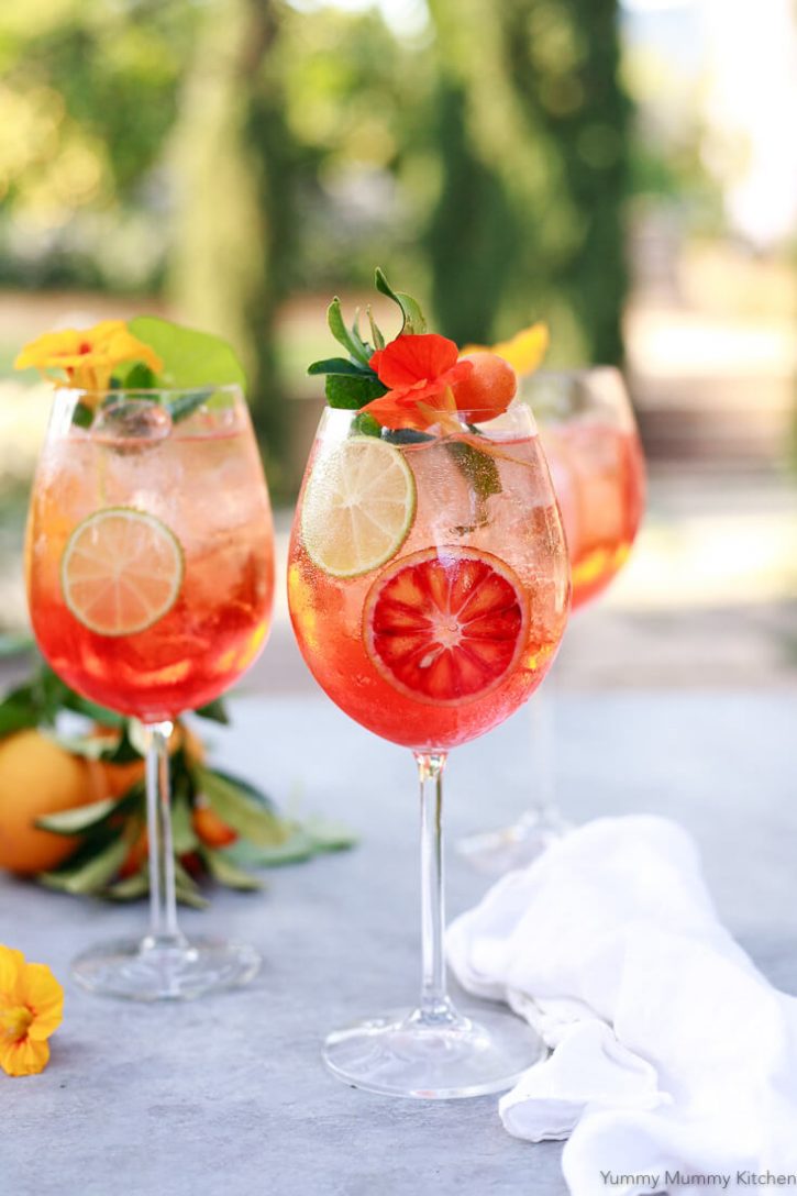 3 wine glasses filled with Aperol Spritz cocktail with blood oranges, limes, and edible flowers sit on a patio table. 