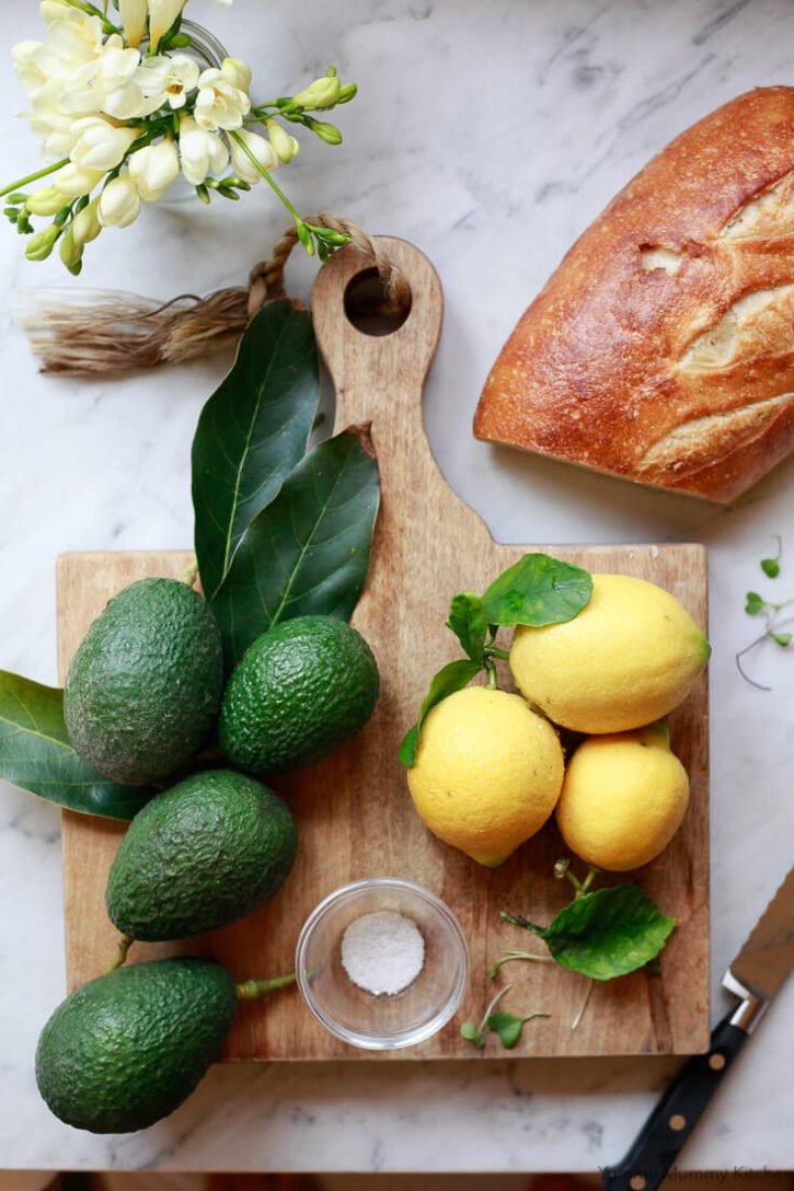 Simple avocado toast ingredients on a cutting board and marble countertop: Haas avocados, lemons, salt, and a loaf of bread. 