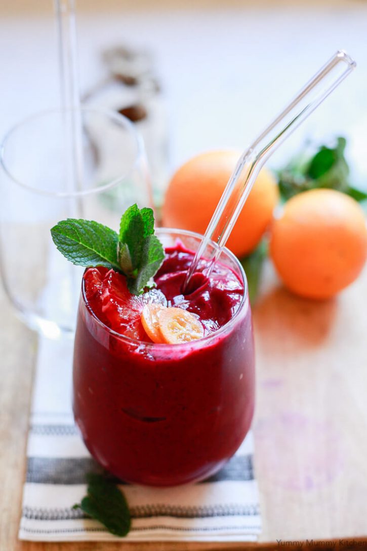 A bright red beet smoothie in a clear glass with a glass straw topped with orange slices and fresh mint. 