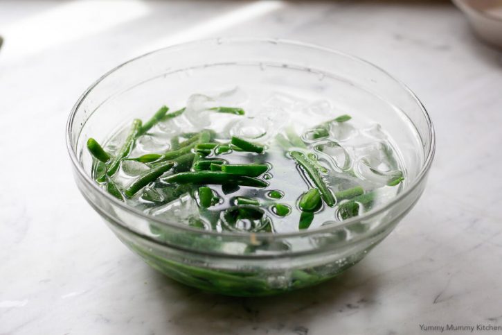 Cooked green beans in a bowl of ice water on a marble counter. 