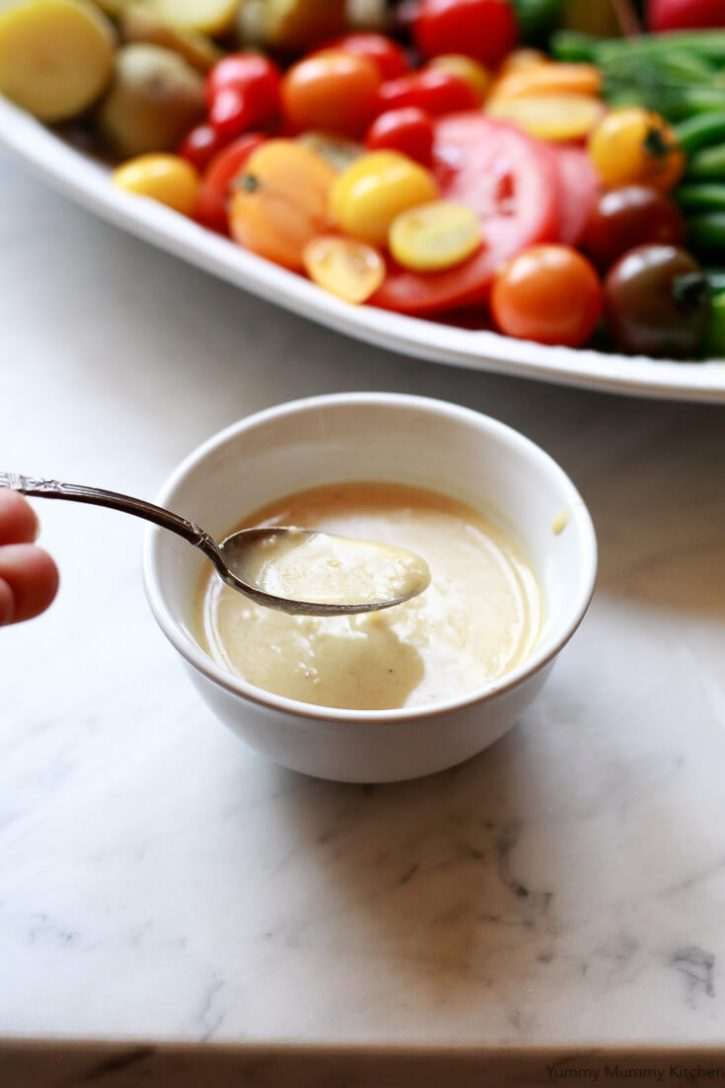 A small white bowl and spoon filled with creamy shallot dijon vinaigrette is the dressing for Nicoise salad. 