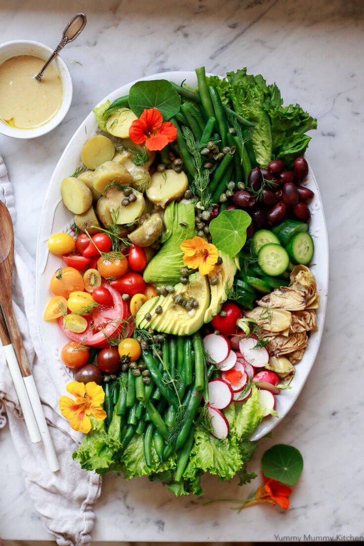 A beautiful Nicoise salad platter with green beans, potatoes, tomatoes, capers, olives, and avocado on a marble countertop with shallot vinaigrette on the side. 