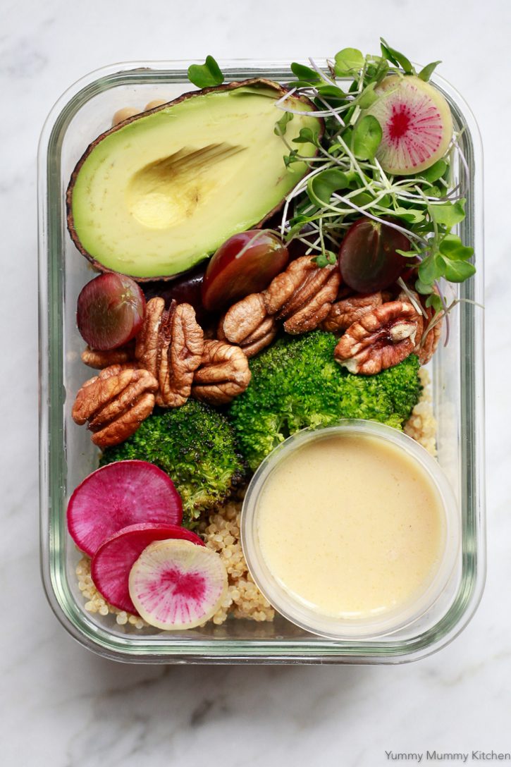 Vegan meal prep lunch or dinner with quinoa, roasted broccoli, pecans, grapes, and avocado. This beautiful meal is in a glass meal prep container with a small container of lemon dressing. 