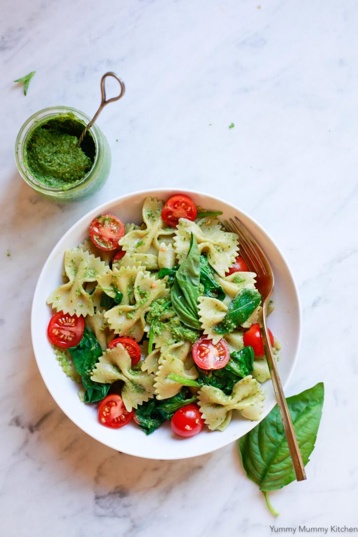 A bowl of vegan pesto pasta sits on a white marble countertop with a small jar of pesto sauce on the side. 