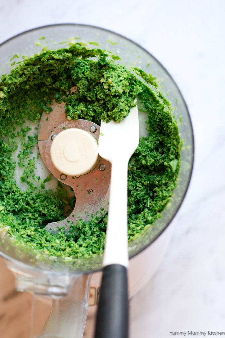A white spatula scrapes down the sides of a food processor while making fresh basil pesto. 
