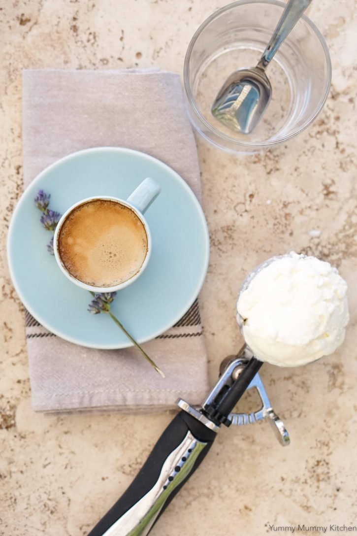 A shot of espresso in a cup, a scoop of vanilla ice cream in an ice cream scooper, and a glass with spoon on a tabletop to make affogato. 