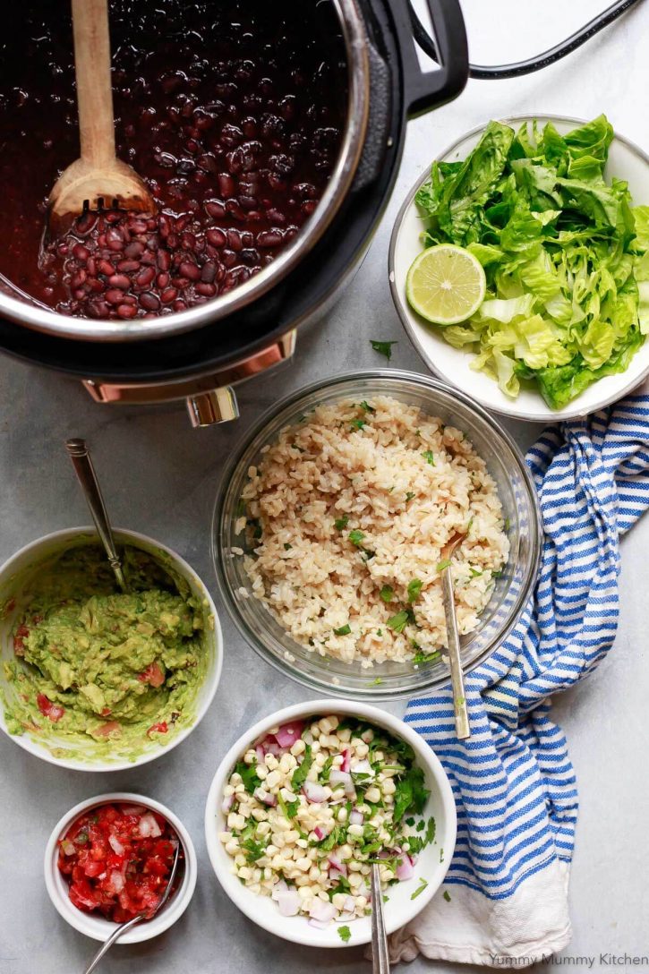 Vegetarian vegan Chipotle burrito bowl toppings sit on a countertop: cilantro lime brown rice, black beans, fresh corn salsa, Romaine lettuce, guacamole, and salsa. 