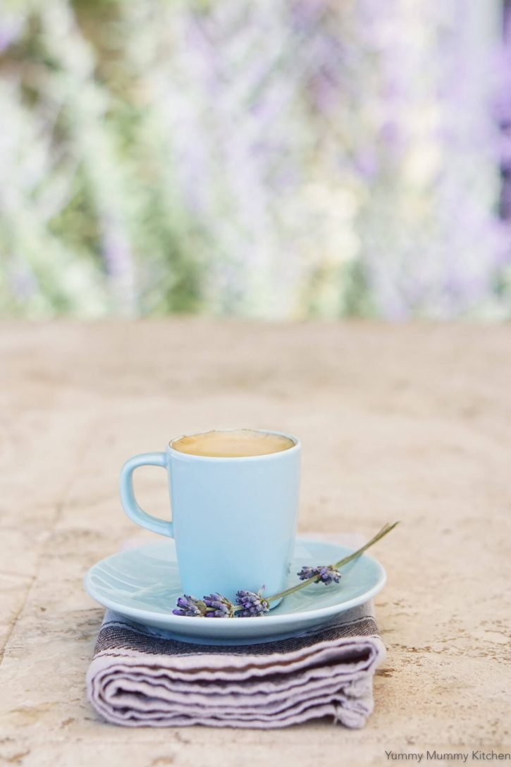 A small blue mug filled with a lavender latte sits on a saucer with fresh lavender sprig. 