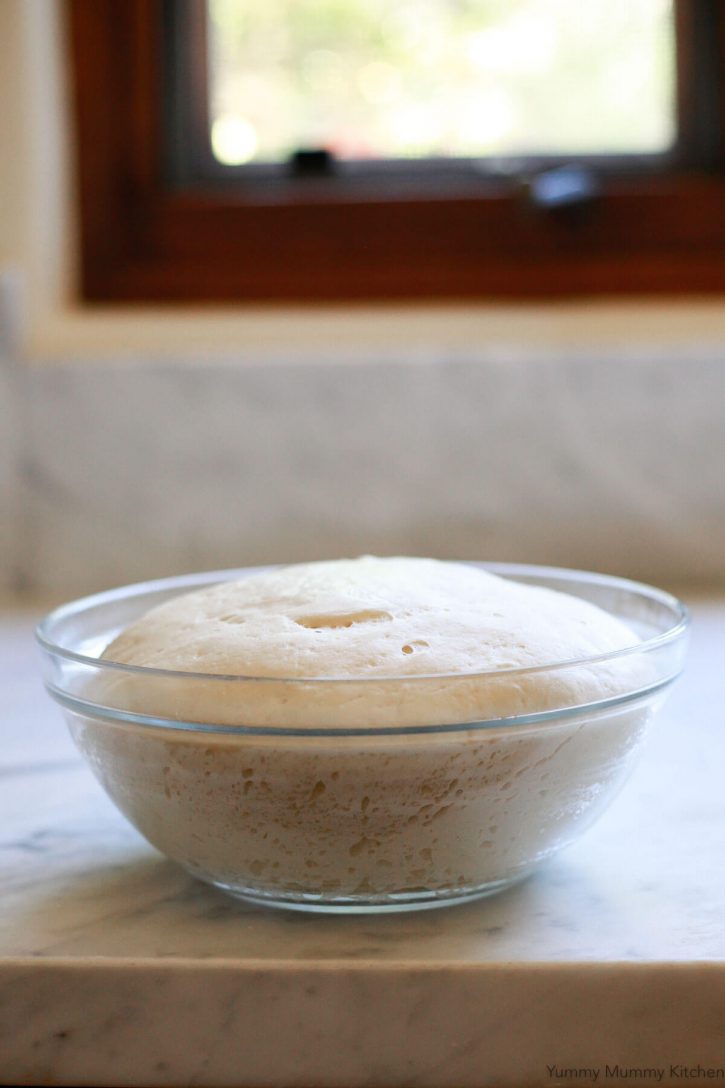 A glass bowl filled with homemade vegan pizza dough sits on a marble countertop. 