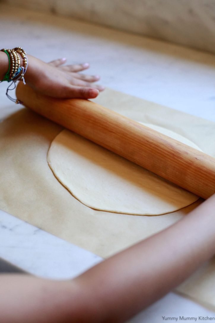 A child's hands roll a thin crust pizza dough with a wooden rolling pin. 