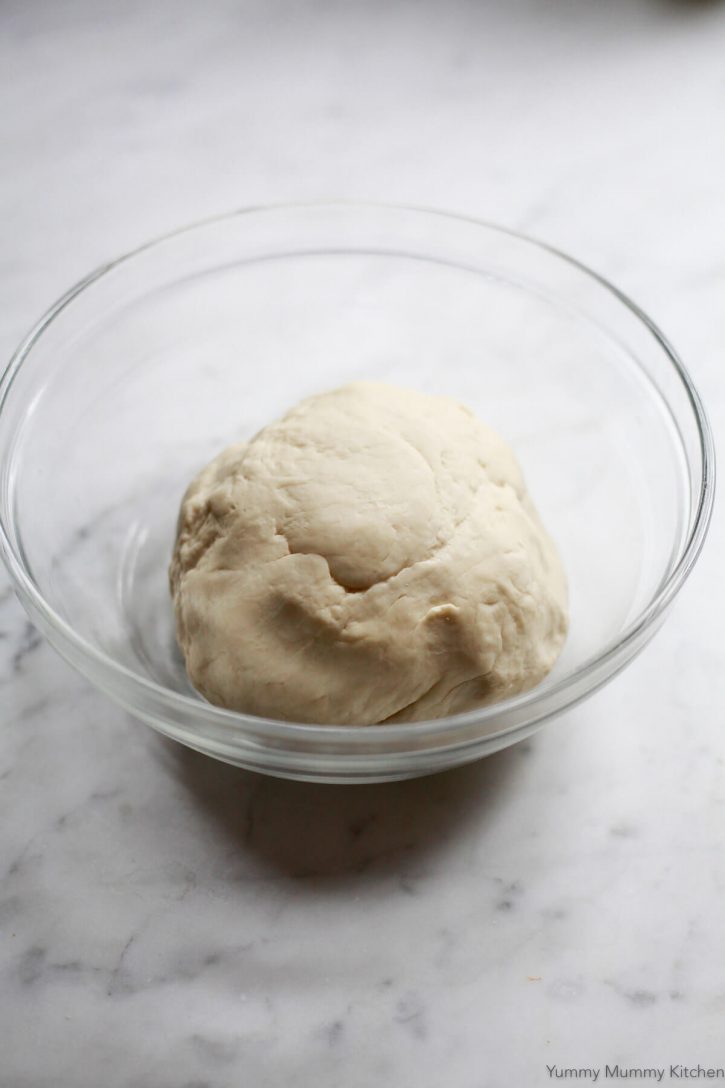 A ball of pizza dough sits in a glass bowl on a marble countertop to begin rising. 