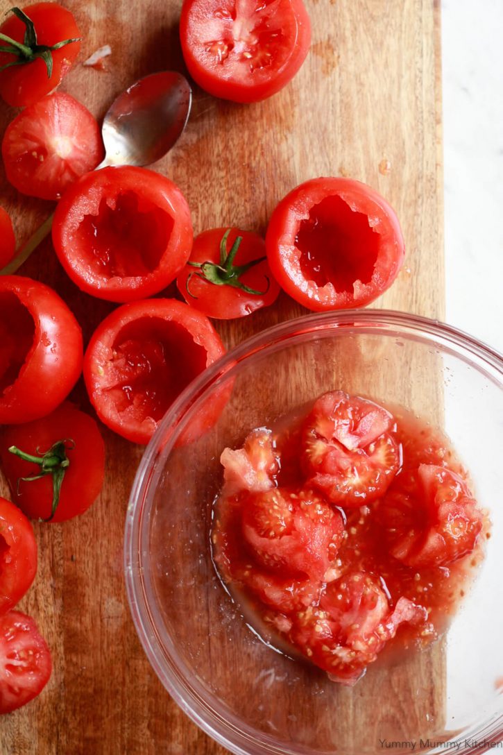 Tomatoes on tops cut off sit on a cutting board with pulp scooped out into a glass bowl. 