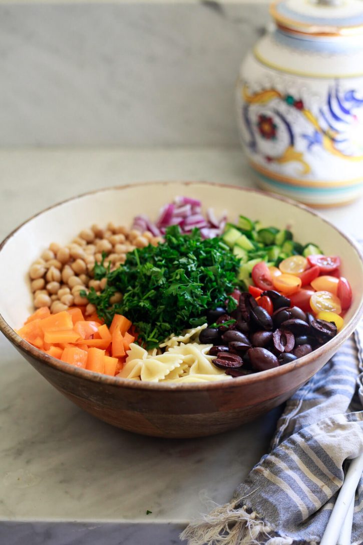 A wooden salad bowl filled with ingredients for a Greek vegan pasta salad. 