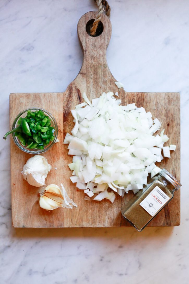 A cutting board with the ingredients for Mexican pinto beans. Chopped onion, cumin, garlic, and jalapeno. 