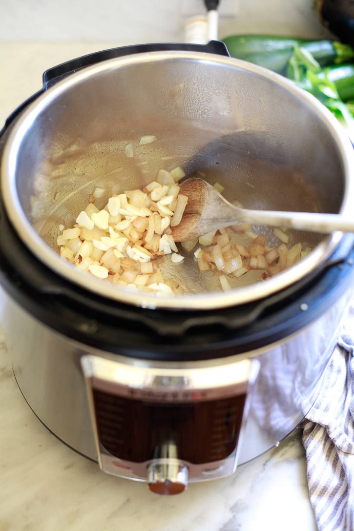 A wooden spoon stirs yellow onions in an Instant Pot on saute mode. 