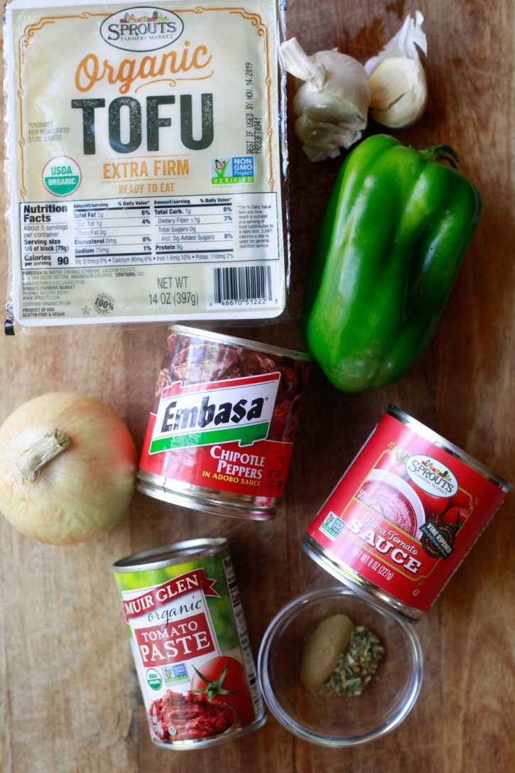 The ingredients for sofritas sit on a wooden board and include tofu, garlic, green bell pepper, tomato sauce, chipotle peppers, tomato paste, oregano and cumin, and a yellow onion. 
