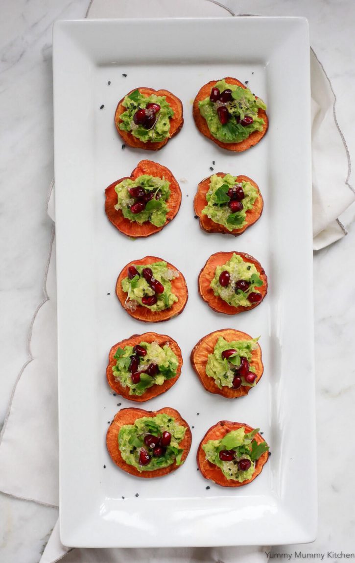 A platter filled with avocado sweet potato toast crostini topped with pomegranate, finger lime, and cilantro. Sweet potato crostini is a healthy vegan appetizer recipe. 