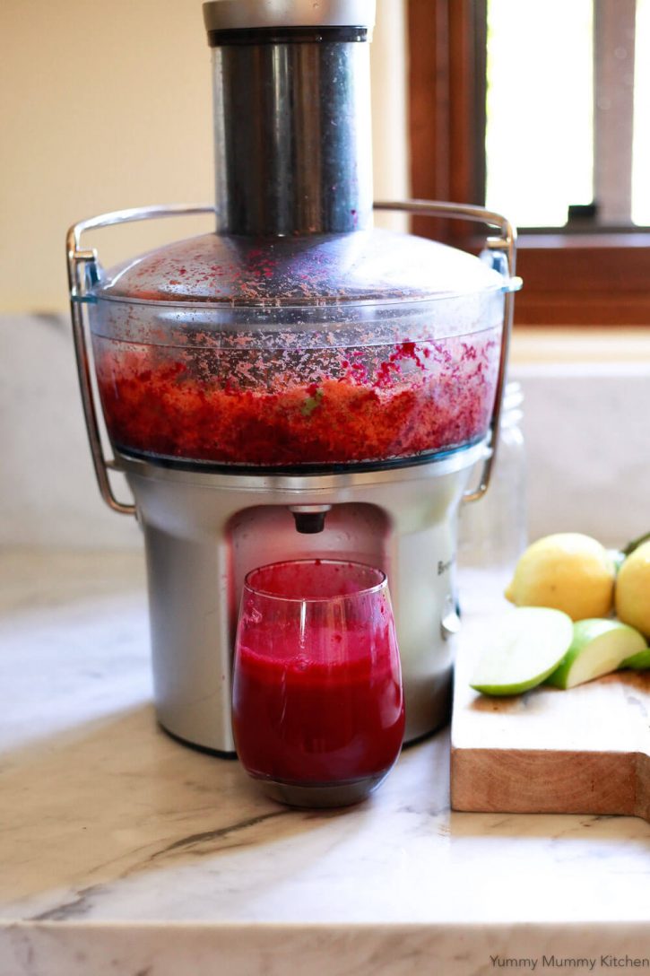 A Breville juicer making a detox beet juice recipe in a white kitchen. A glass of bright red beet juice below the juicer. 